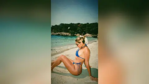 Blonde woman in a blue bikini relaxing on a sunny tropical beach by turquoise ocean water.