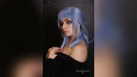 Woman with long lavender wig and black off-the-shoulder outfit posing in a moody studio setting.