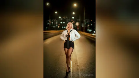 Confident blonde woman in a white shirt and black mini skirt posing on a city street at night under glowing lights.