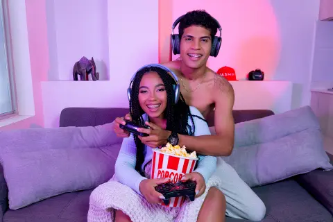 Happy young couple in casual loungewear enjoying popcorn and gaming together on a cozy sofa with neon lighting.