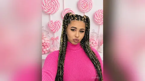 Young woman with long blonde braids wearing a pink turtleneck sweater in front of pink heart decorations.