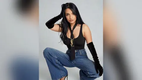 Stylish woman in black tank top, YSL tie, and long velvet gloves posing in blue jeans against a white background.