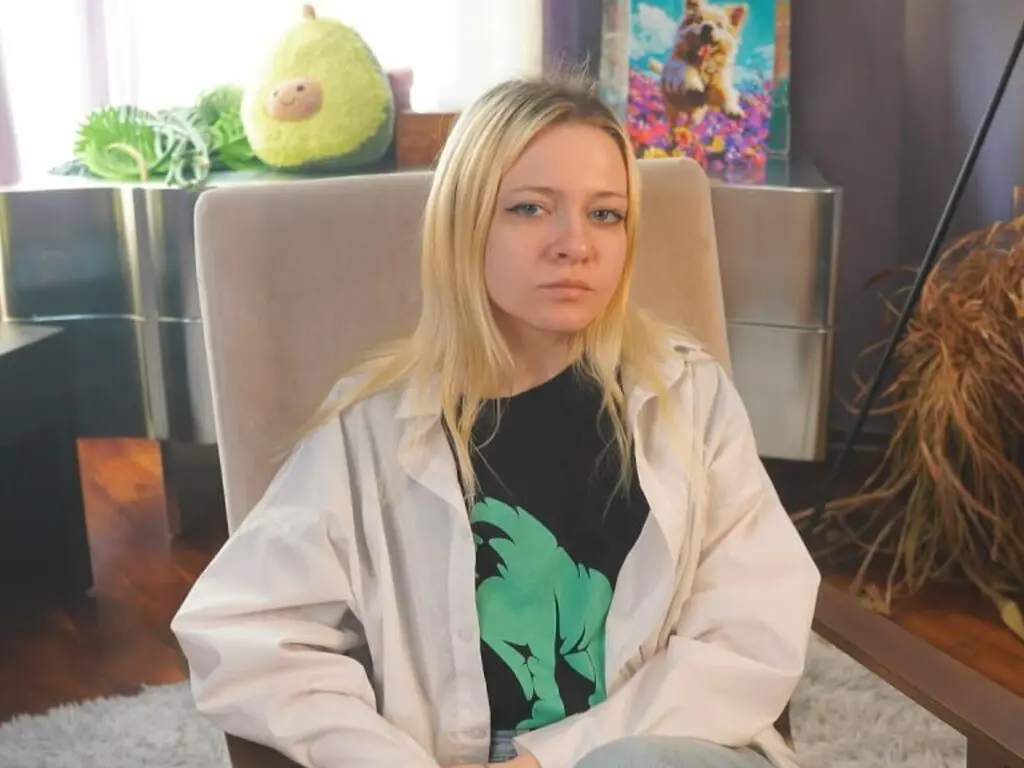 Blonde woman in a black graphic t-shirt and white shirt sitting on a chair in a cozy living room.