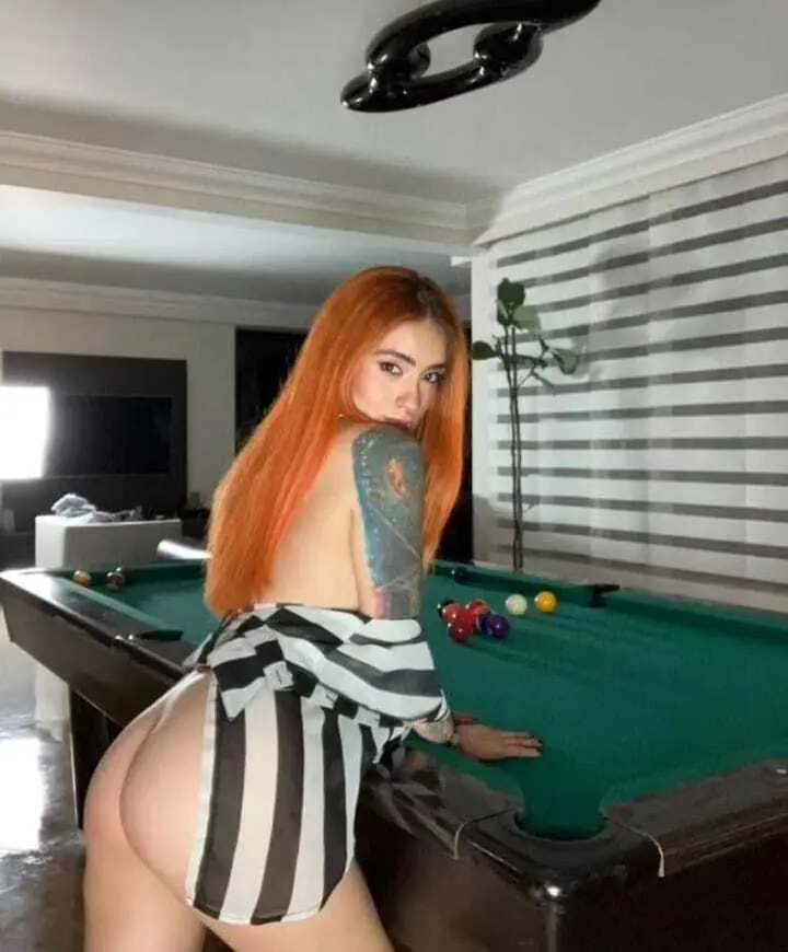 IsabellaVillalobos has long, wavy orange hair and looks directly at the camera while lying on a white bedsheet.