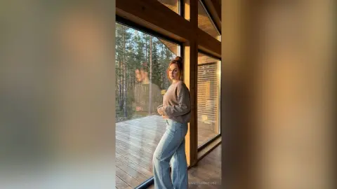 Redheaded woman in a brown sweater and light blue jeans standing by a large window in a cozy wooden cabin.