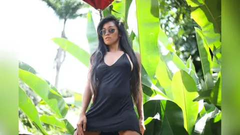MokaAkashiya — Stylish woman in a black sleeveless dress posing amidst lush green tropical banana leaves in bright sunlight.