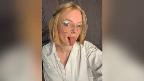 Blonde woman in a white button-down shirt wearing clear glasses and making a playful expression indoors.