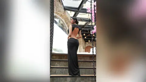 Aleja stands on stone stairs wearing a long black backless dress that shows her bare back and curves.
