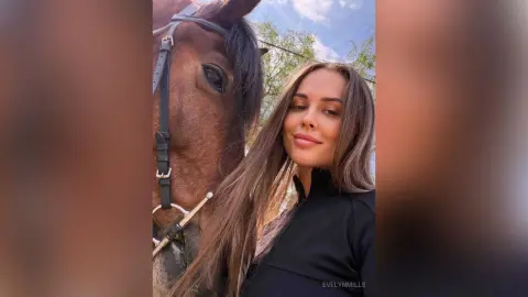 EvelynMills smiles next to a brown horse while wearing a black long-sleeved top outdoors.