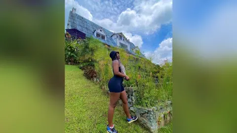 ValeryMei stands outdoors in a black bodysuit, posing against a lush green garden and a large house under a blue sky.