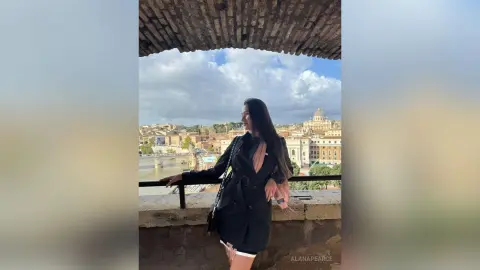 Stylish woman in a black coat standing on a stone balcony overlooking a historic European city under a cloudy sky.