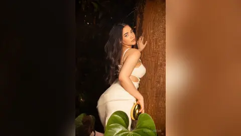 Elegant woman in a white cocktail dress posing against a large tree trunk in a moody, natural outdoor setting.