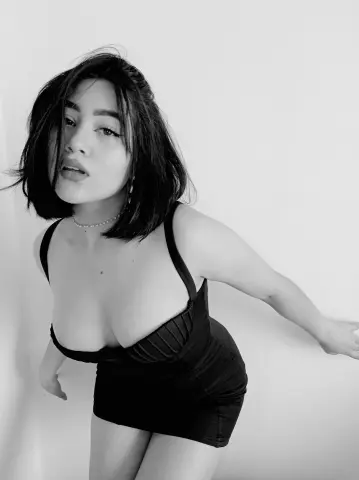 AnnaKomatsuu poses in a black low-cut dress against a plain white wall, looking toward the camera with a slight pout.