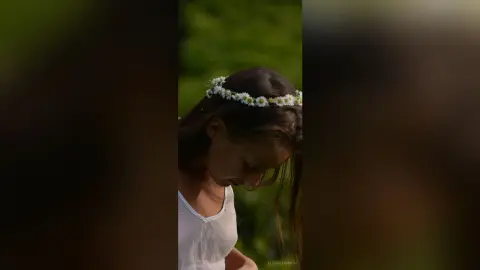 Dreamy portrait of a girl in a white dress wearing a daisy flower crown in a lush green field.