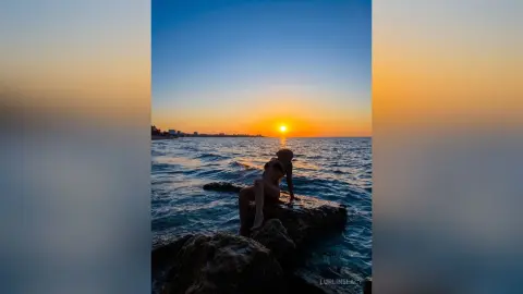 LurlineLacy sits on rocks in the ocean at sunset, wearing a black bikini.