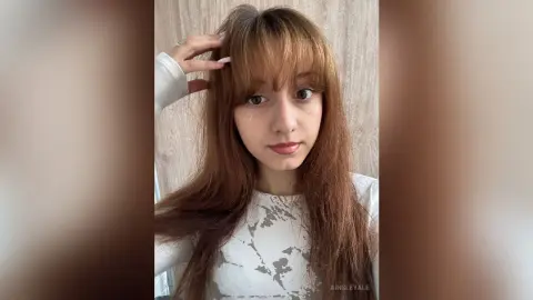 Young woman with long brown hair and glitter makeup wearing a white patterned top in a bright indoor setting.