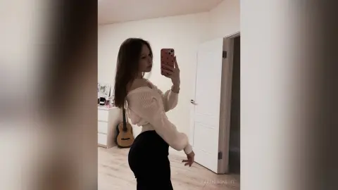 TarenLeonetti poses in a white long-sleeve sweater and black pants, holding a phone for a mirror selfie in a room with a guitar.