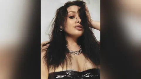 Glamorous woman with long dark hair wearing a black strapless top and silver chain necklace in a moody setting.