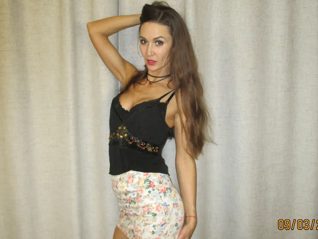 Young woman in a black lace camisole and floral skirt posing against a neutral curtain backdrop.