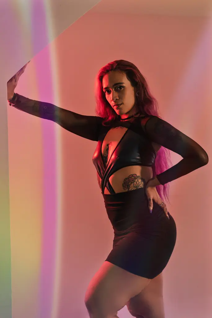 Tattooed woman in black lace gloves and choker posing with a moody expression against red curtains.