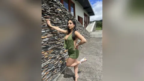 Glamorous woman in a green sequin mini dress posing by a textured stone wall on a sunny day.