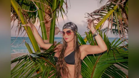 AndreaKalo poses in a black bikini top and sunglasses, framed by large green palm leaves on a beach at sunset.
