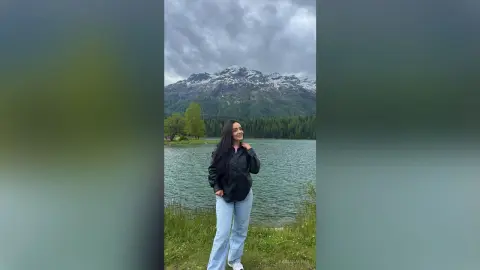 FabianaLima stands in a grassy field by a lake with snow-capped mountains in the background, wearing a black button-down shirt and light blue jeans.
