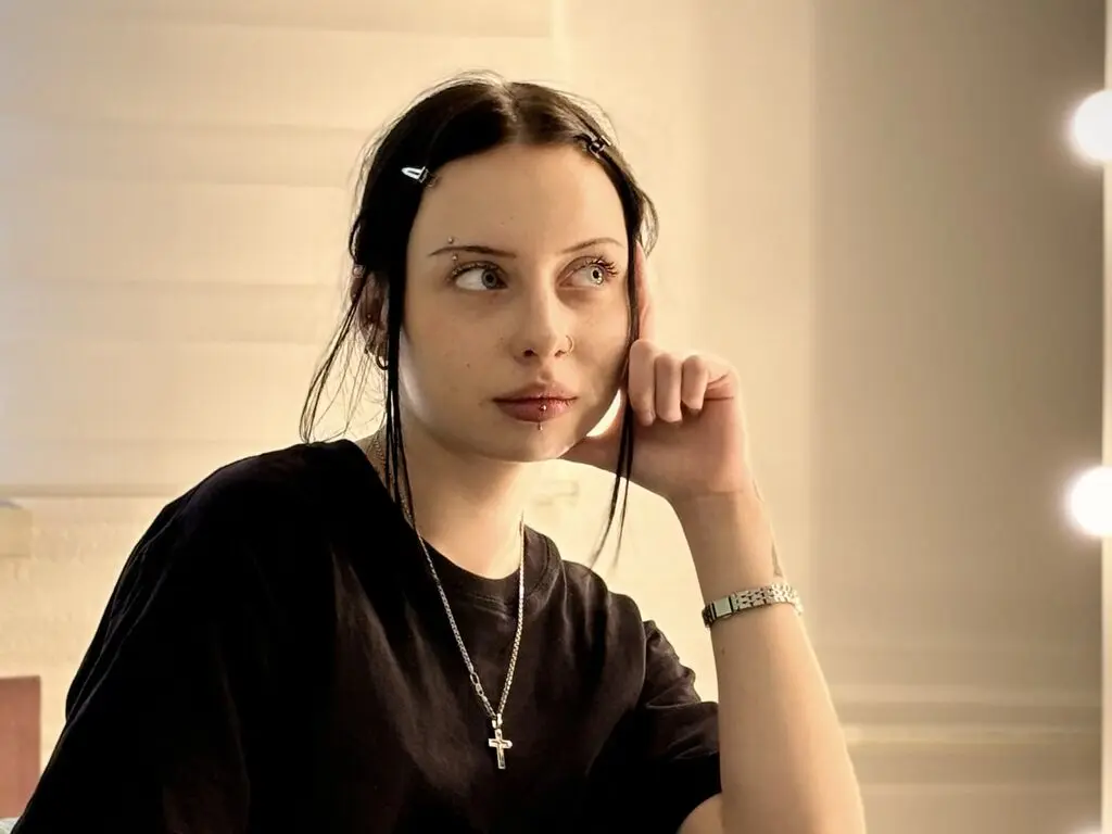 Young woman with dark hair and silver cross necklace looking away in a soft, moody indoor setting.