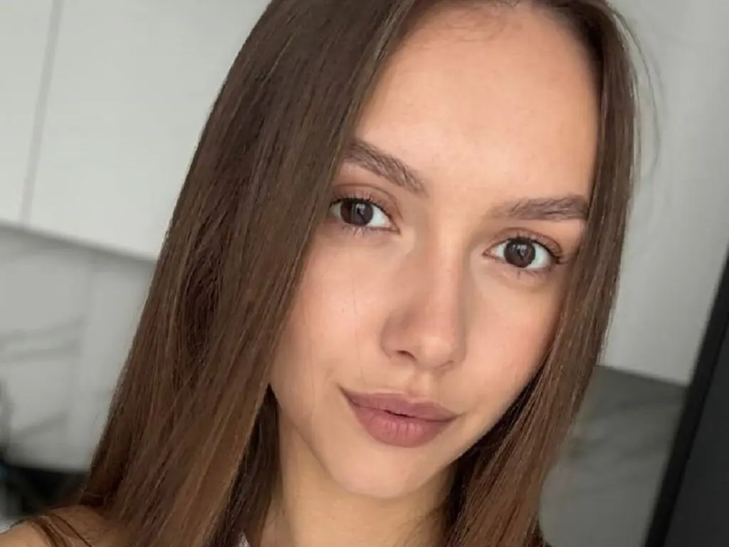 Close-up portrait of a young woman with long brown hair and natural makeup in a bright indoor setting.