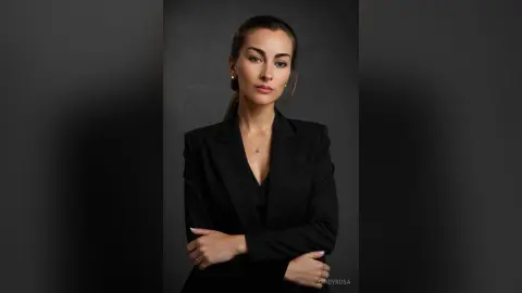 Confident professional woman in a black blazer and necklace posing against a dark studio background.