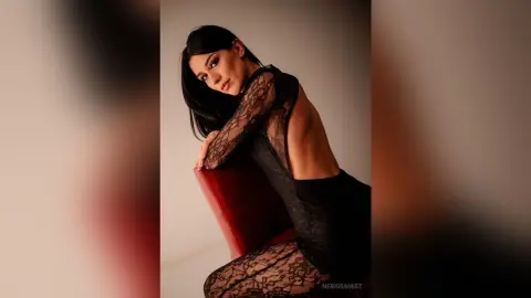 NerissaMist poses in a black lace bodysuit while leaning over a red chair with her long dark hair down.