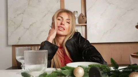 Blonde woman in a black velvet outfit relaxing at a dining table with festive greenery and glassware.