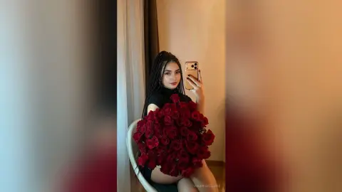 Young woman with long braids in a black turtleneck holding a large bouquet of red roses for a romantic selfie.