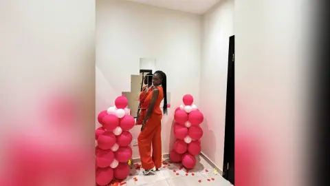 Stylish Black woman in a red jumpsuit posing next to pink balloon decor in a modern white room.