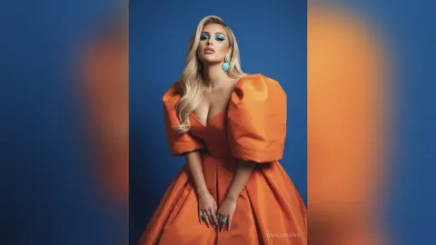 Glamorous blonde woman in an elegant orange puff-sleeve gown posing against a blue studio background.