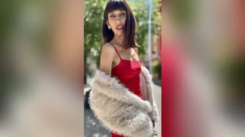 Young woman in a red dress and fluffy faux fur coat posing outdoors in soft sunlight.