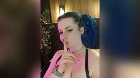 Young woman with blue hair in a black halter top posing thoughtfully in a dimly lit restaurant setting.