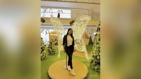 Woman in black trousers and white top standing by a large glowing butterfly decoration in a festive indoor setting.