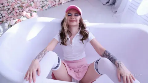 AmeliePearly wearing a white polo shirt and pink skirt, sitting in a white bathtub with her long hair down and a pink cap on.