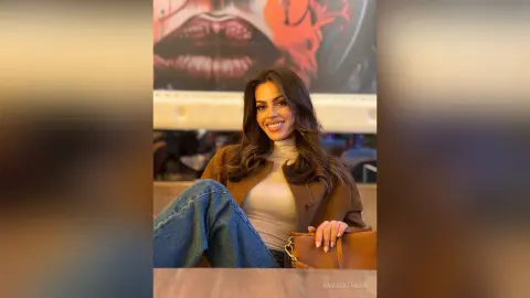 Smiling brunette woman in a beige turtleneck and brown jacket posing in front of urban street art.