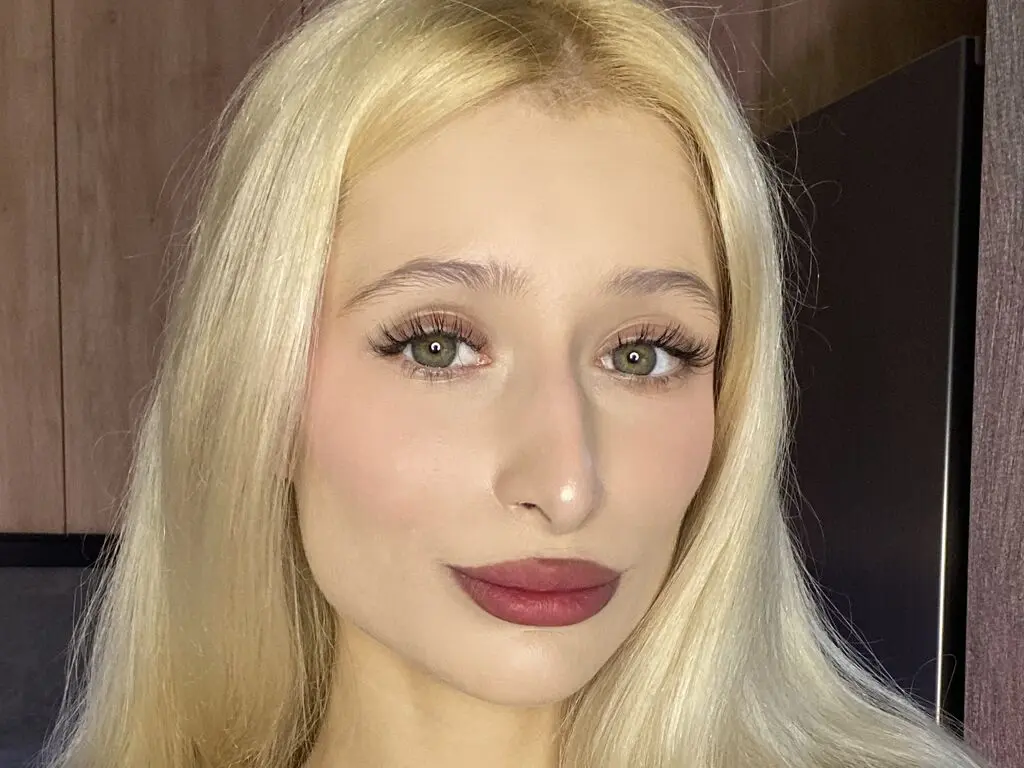 Close-up portrait of a blonde woman with green eyes and natural makeup in a bright indoor setting.