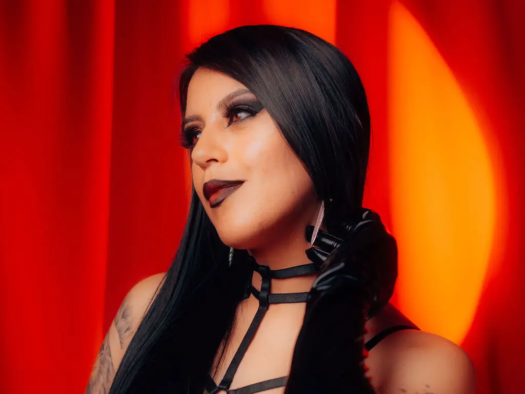 Woman with dark hair and gothic makeup wearing a black strap outfit against a vibrant red curtain background.