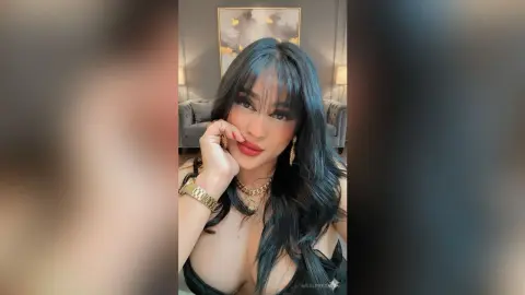 Young woman with long dark hair wearing gold jewelry and a black top in a cozy living room setting.