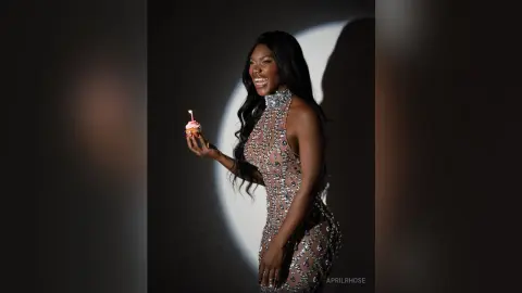 Smiling woman in a sparkling sequin halter dress holding a cupcake against a dark studio background.