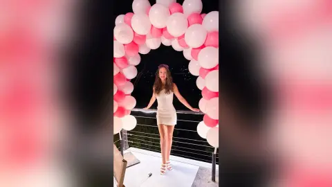 Smiling young woman in a white mini dress posing under a pink and white balloon arch on a balcony.