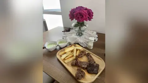 Steak and french fries served on a wooden platter with dipping sauce and pink roses on a dining table.