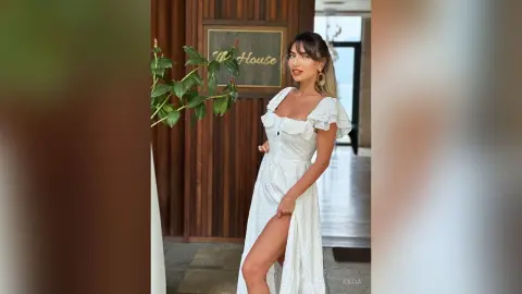 Katia poses in a white sundress with a high slit, showing off her long legs and brunette hair in a modern indoor setting.