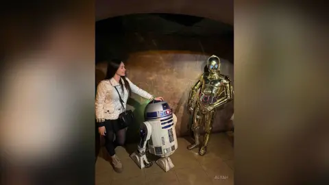Woman in white jacket standing next to R2-D2 and C-3PO droid statues in a dimly lit museum setting.