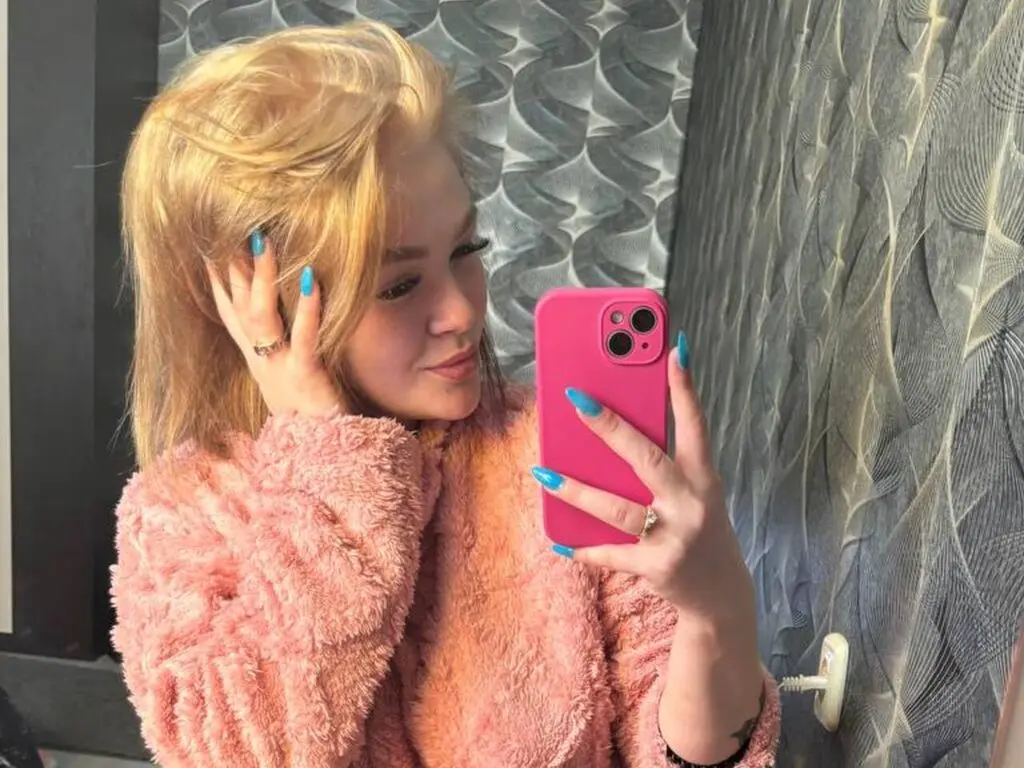 ElizabethCamaro poses for a mirror selfie wearing a pink fuzzy sweater, showing off her blonde hair and bright blue fingernails against a textured grey wall.