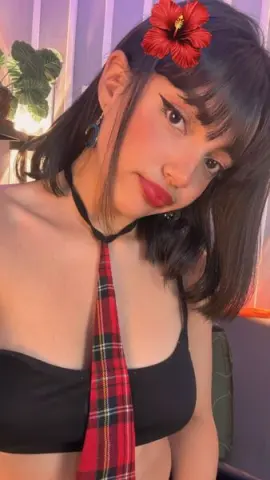 Young woman with a red hibiscus flower in her hair wearing a black crop top and red plaid tie in a cozy room.